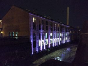 A building at nighttime. It is located along an overgrown canal. The side of the building is lit with purple columns of light. In the distance, a white smokestack is visible.