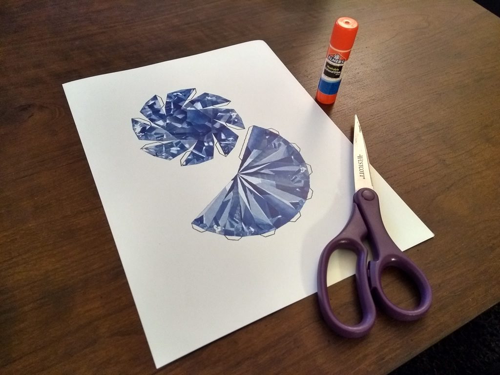 A printout of a flat model of a blue sapphire, sitting on a table. Next to the paper are a pair of scissors and a glue stick.