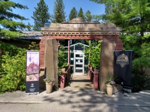 The entrance to Cyan Worlds headquarters in Mead, WA. A red stone arch stands in front of a small walkway bridge to the front door. On other side of the entrance are banners for the games Obduction and Firmament, with conifer trees further to the sides.